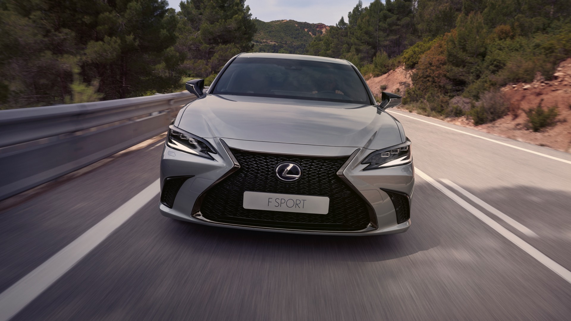 The front of a Lexus ES F Sport as it speeds along a highway. Its headlights are on.