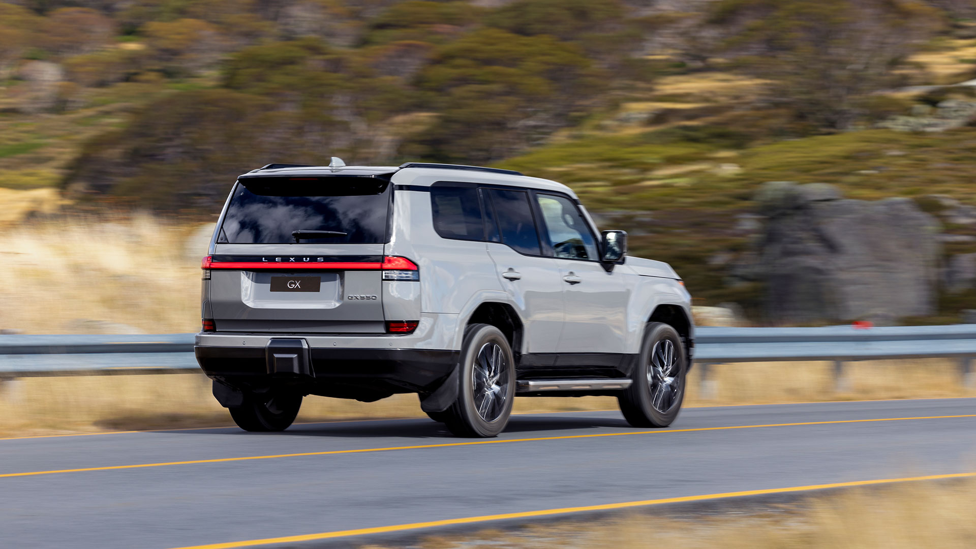 A grey Lexus LX drives along a road with greenery in the background